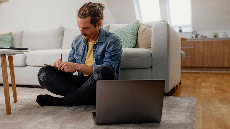 Homem jovem, sentado no chão da sala, encostado a um sofá, a tirar notas num caderno em cima do joelho e com um computador portátil aberto, em cima de um tapete felpudo
